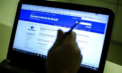 Pessoa segura uma caneca em frete um notebook aberto com a página da Receita Federal ao fundo