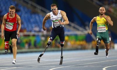Três atletas amputados disputam a prova dos 100 m: um britânico, de branco, que lidera em uma pista central, um dinamarquês de vermelho, à esquerda, e um australiano, de amarelo, à direita