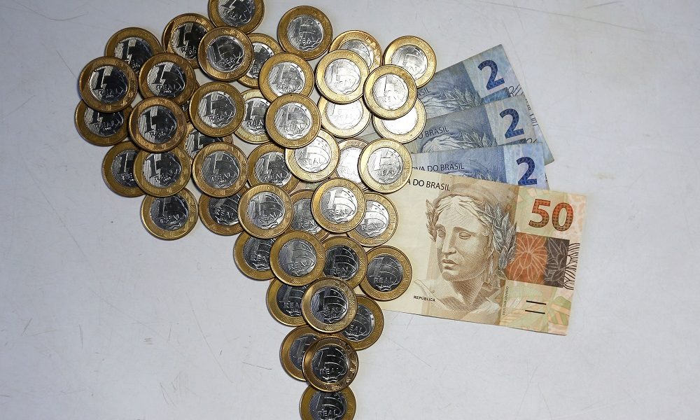 Moedas de R$ 1 formam o mapa do Brasil; do lado direito, há três notas de R$ 2 e uma de R$ 50